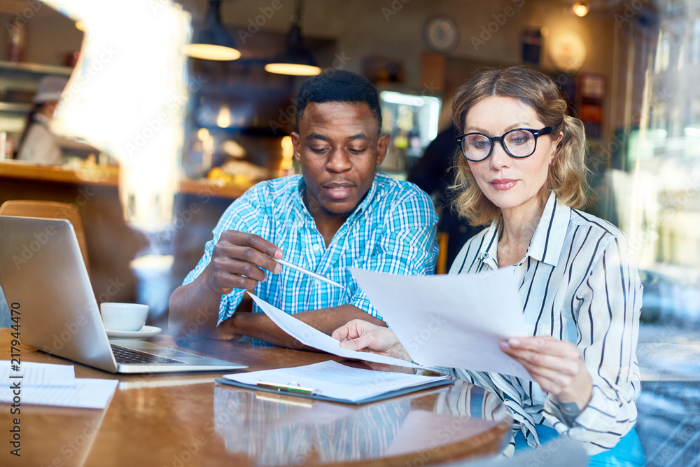 African-American man and woman in glasses sitting at table and reading documents together during business meeting in cafe African-American man and woman in glasses sitting at table and reading documents together during business meeting in cafe