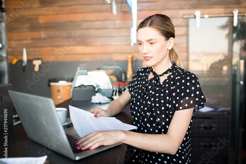 Stylish young woman using laptop and holding papers standing at counter in shop managing business 