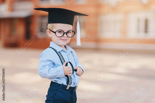 Little smart boy in spectacles, academic hat and glasses standing in the backyard of the school. Holds a magnifying glass in his hands