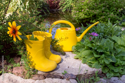 Obraz na plátně Yellow children's rubber boots with flower and yellow garden watering can on a b