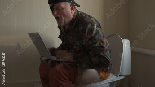 Man with gray hair, beard and mustache in military jacket with laptop sitting on toilet. Man's eye is covered with black medical patch. Dude is infatuated with computer and actively expressed emotions photo