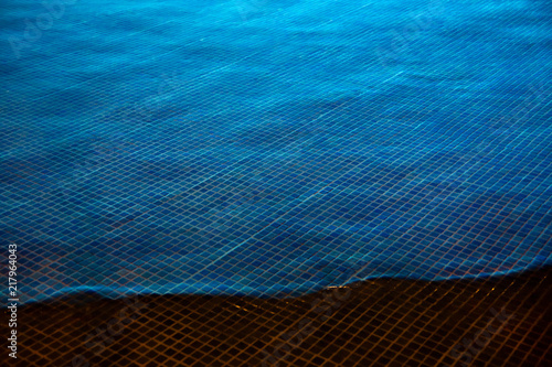 Blue lighted swimming pool water whasing up the pool rim with small square mosaic tiles photo