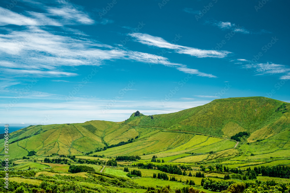 A mountain with green fields and a blue sky with some clouds