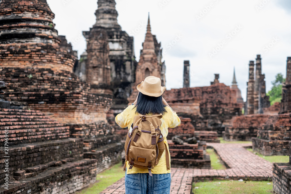 Fototapeta premium Asian tourist woman take a photo of ancient of pagoda temple thai architecture at Sukhothai Historical Park,Thailand. Female traveler in casual thai cloths style visiting city concept.