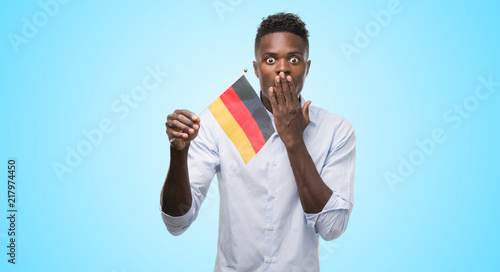 Young african american man holding german flag cover mouth with hand shocked with shame for mistake, expression of fear, scared in silence, secret concept