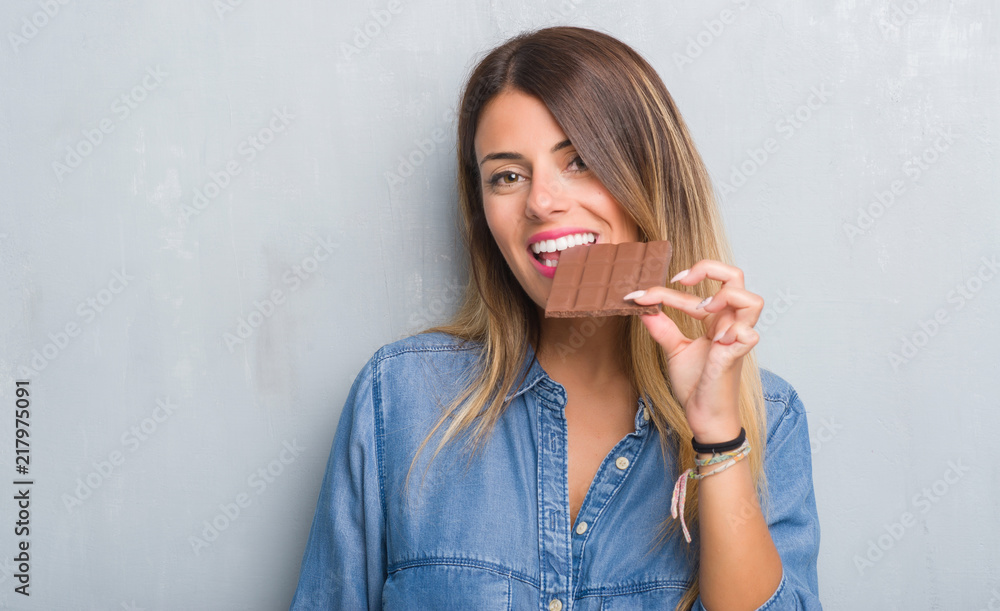 Young adult woman over grey grunge wall eating chocolate bar with a confident expression on smart face thinking serious Young adult woman over grey grunge wall eating chocolate bar with a confident expression on smart face thinking serious