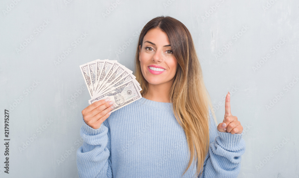 Young adult woman over grey grunge wall holding dollars surprised with an idea or question pointing finger with happy face, number one Young adult woman over grey grunge wall holding dollars surprised with an idea or question pointing finger with happy face, number one