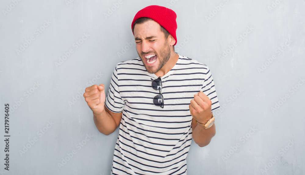 Naklejka premium Handsome young hipster man over grey grunge wall wearing navy t-shirt and wool cap very happy and excited doing winner gesture with arms raised, smiling and screaming for success. Celebration concept.