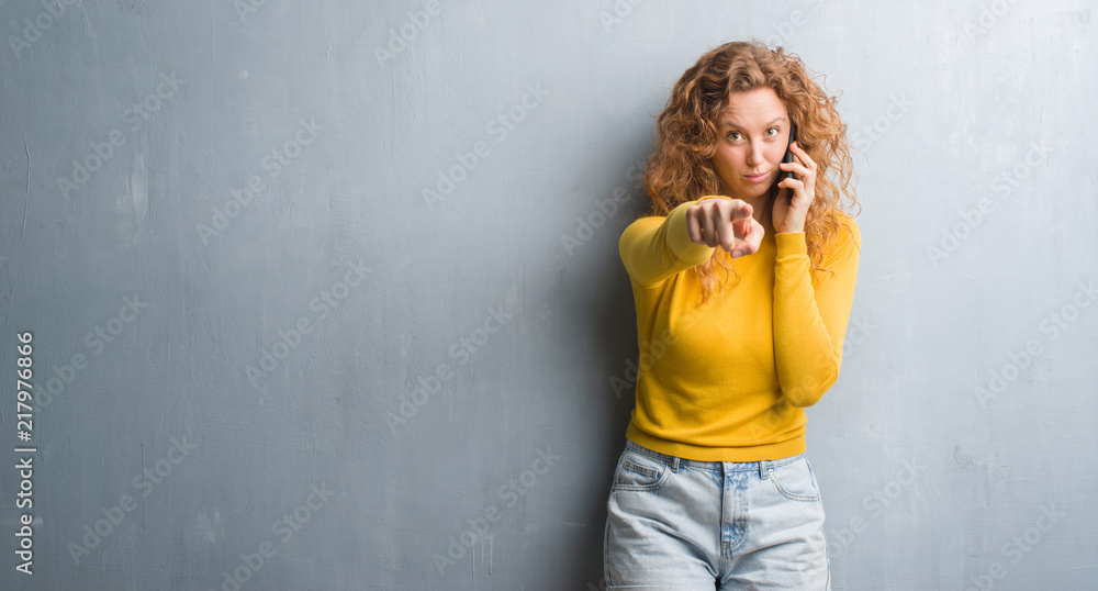Young redhead woman over grey grunge wall talking on the phone pointing with finger to the camera and to you, hand sign, positive and confident gesture from the front