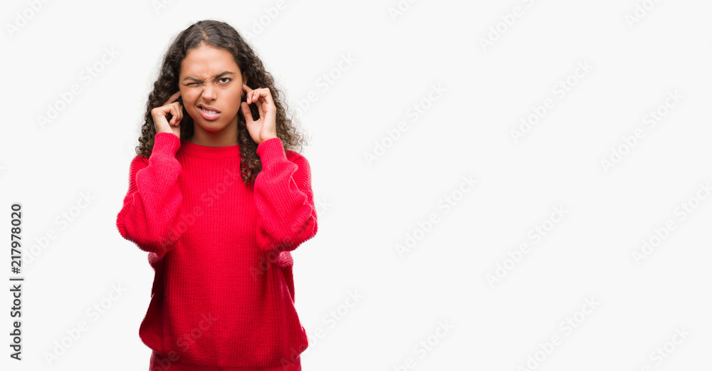 Young hispanic woman wearing red sweater covering ears with fingers with annoyed expression for the noise of loud music. Deaf concept.