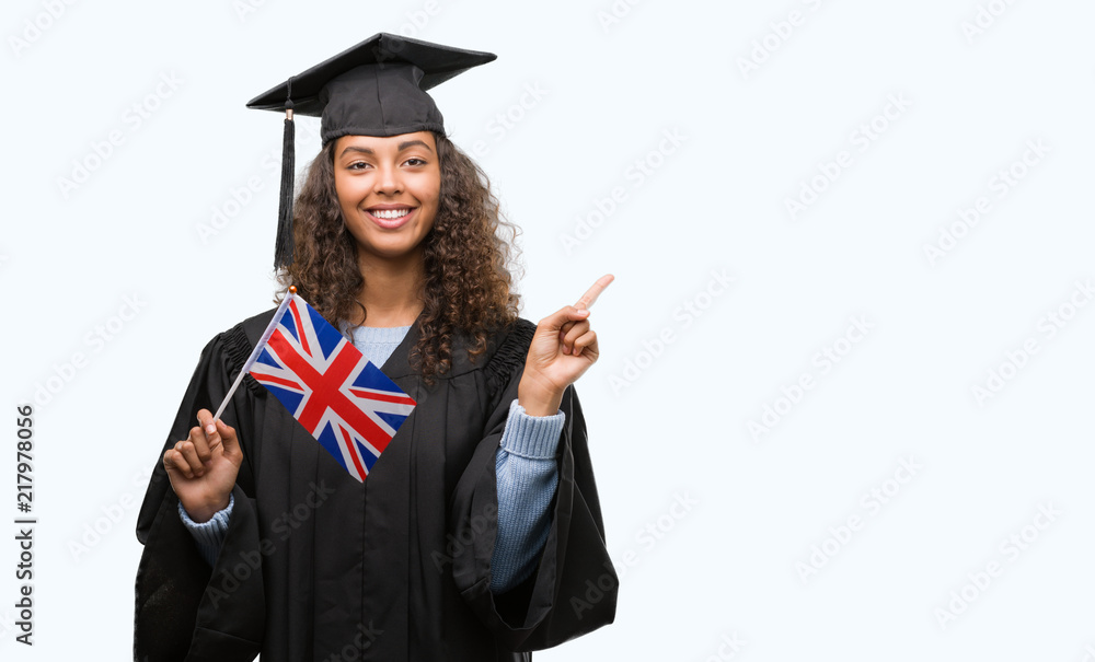 Young hispanic woman wearing graduation uniform holding flag of UK very happy pointing with hand and finger to the side Young hispanic woman wearing graduation uniform holding flag of UK very happy pointing with hand and finger to the side
