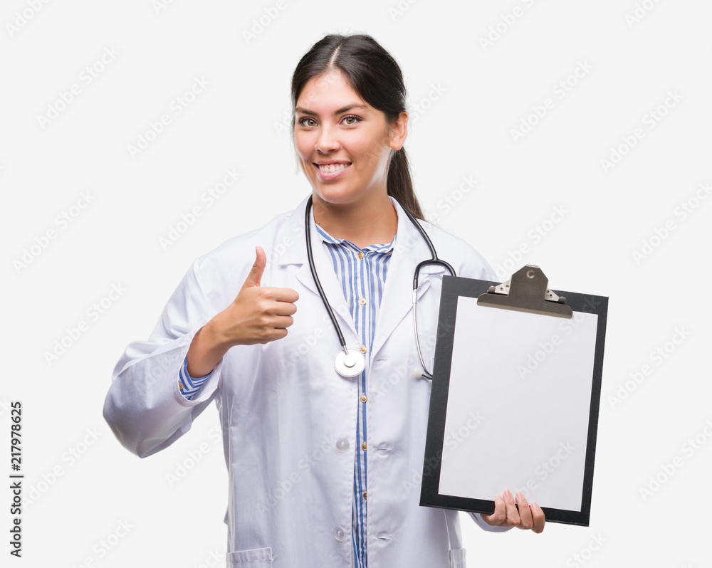Young hispanic doctor woman holding clipboard happy with big smile doing ok sign, thumb up with fingers, excellent sign Young hispanic doctor woman holding clipboard happy with big smile doing ok sign, thumb up with fingers, excellent sign
