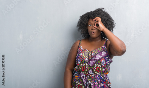 Young african american woman over grey grunge wall wearing colorful dress doing ok gesture shocked with surprised face, eye looking through fingers. Unbelieving expression.