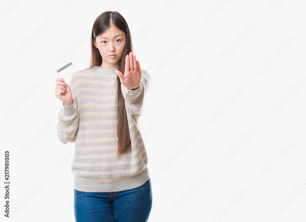 Young Chinese woman over isolated background holding credit card with open hand doing stop sign with serious and confident expression, defense gesture Young Chinese woman over isolated background holding credit card with open hand doing stop sign with serious and confident expression, defense gesture