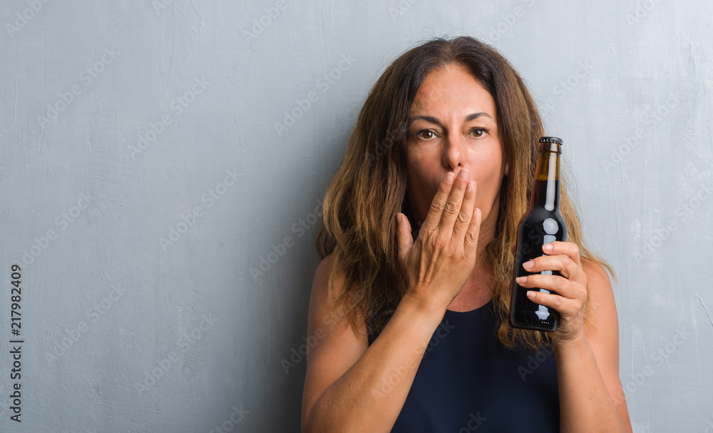 Middle age hispanic woman standing over grey grunge wall holding beer bottle cover mouth with hand shocked with shame for mistake, expression of fear, scared in silence, secret concept Middle age hispanic woman standing over grey grunge wall holding beer bottle cover mouth with hand shocked with shame for mistake, expression of fear, scared in silence, secret concept