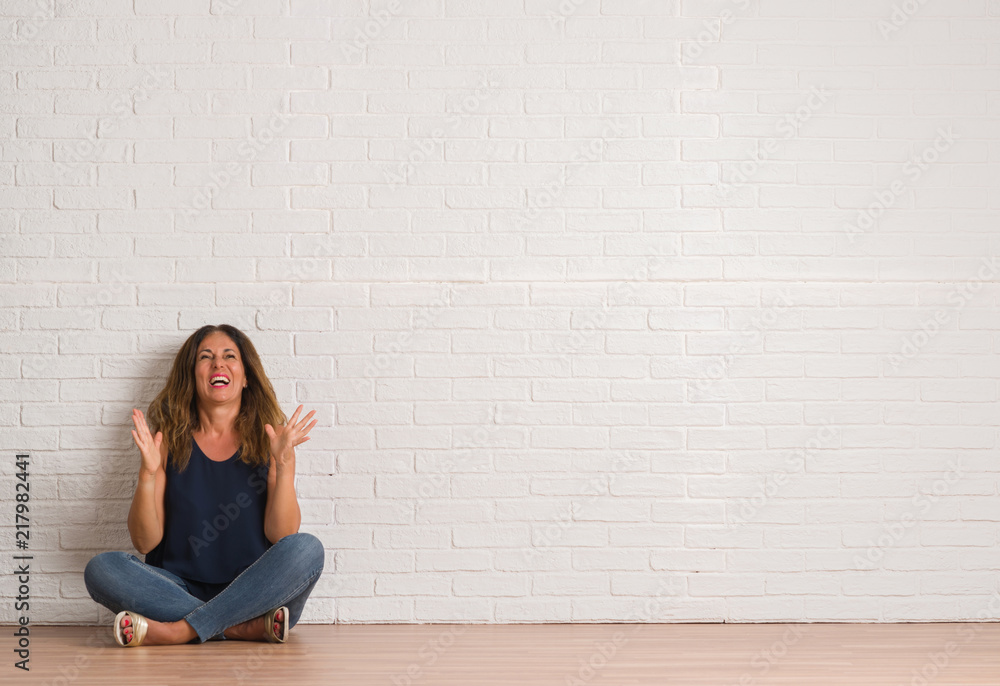 Middle age hispanic woman sitting on the floor over white brick wall crazy and mad shouting and yelling with aggressive expression and arms raised. Frustration concept. Middle age hispanic woman sitting on the floor over white brick wall crazy and mad shouting and yelling with aggressive expression and arms raised. Frustration concept.
