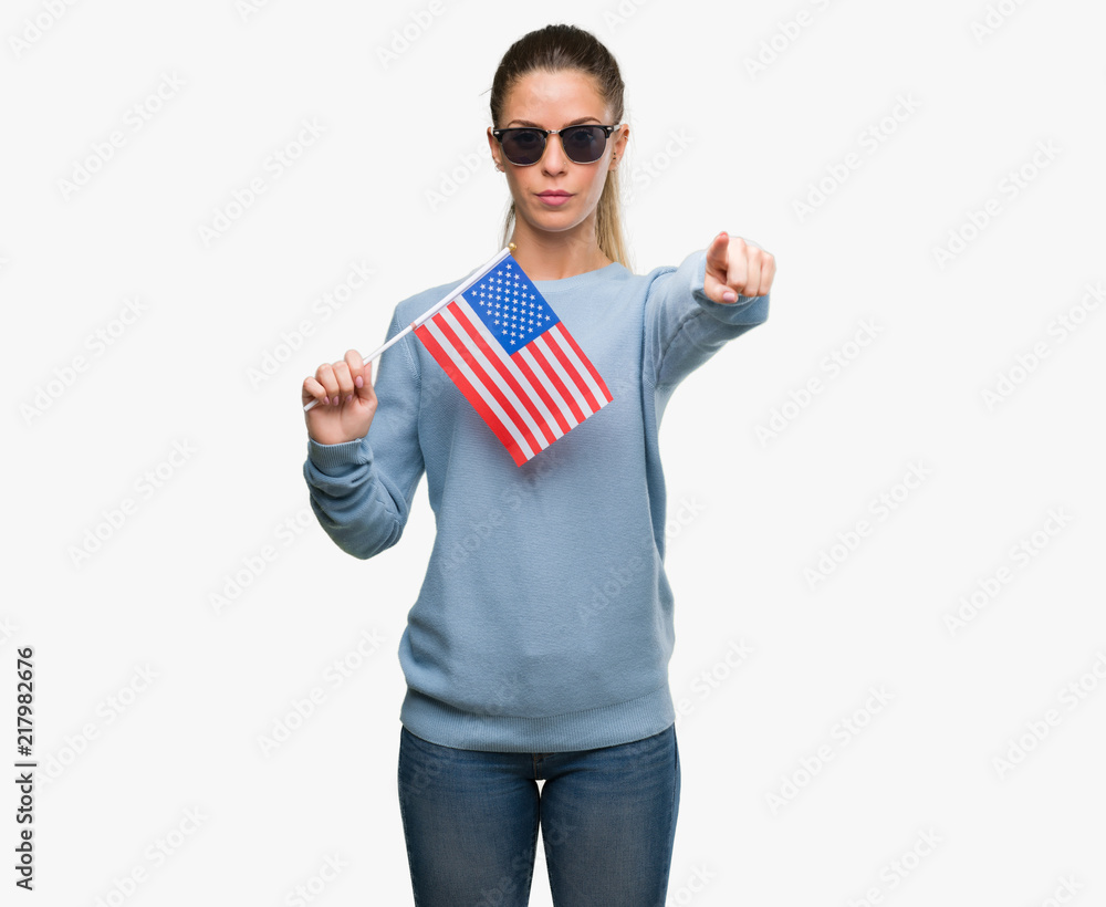Beautiful young woman holding USA flag pointing with finger to the camera and to you, hand sign, positive and confident gesture from the front Beautiful young woman holding USA flag pointing with finger to the camera and to you, hand sign, positive and confident gesture from the front