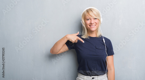 Adult caucasian woman over grunge grey wall wearing headphones with surprise face pointing finger to himself