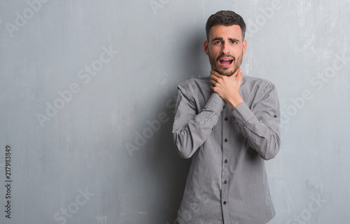 Young adult man standing over grey grunge wall shouting and suffocate because painful strangle. Health problem. Asphyxiate and suicide concept.