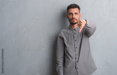 Young adult man standing over grey grunge wall looking unhappy and angry showing rejection and negative with thumbs down gesture. Bad expression.