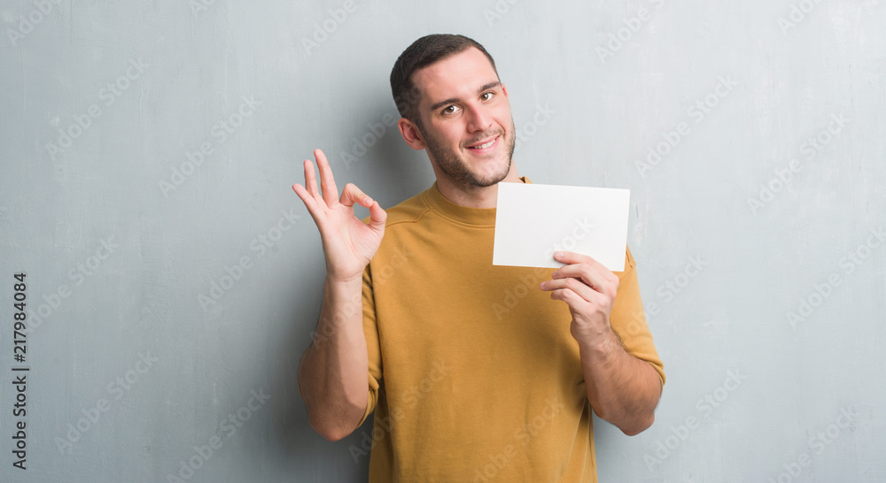 Young caucasian man over grey grunge wall holding blank card doing ok sign with fingers, excellent symbol Young caucasian man over grey grunge wall holding blank card doing ok sign with fingers, excellent symbol