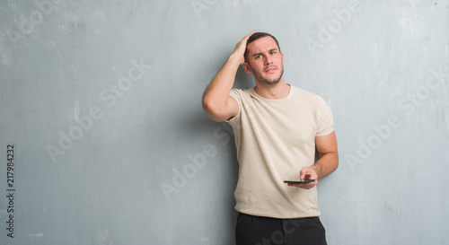 Young caucasian man over grey grunge wall texting a message using smartphone stressed with hand on head, shocked with shame and surprise face, angry and frustrated. Fear and upset for mistake. © Krakenimages.com