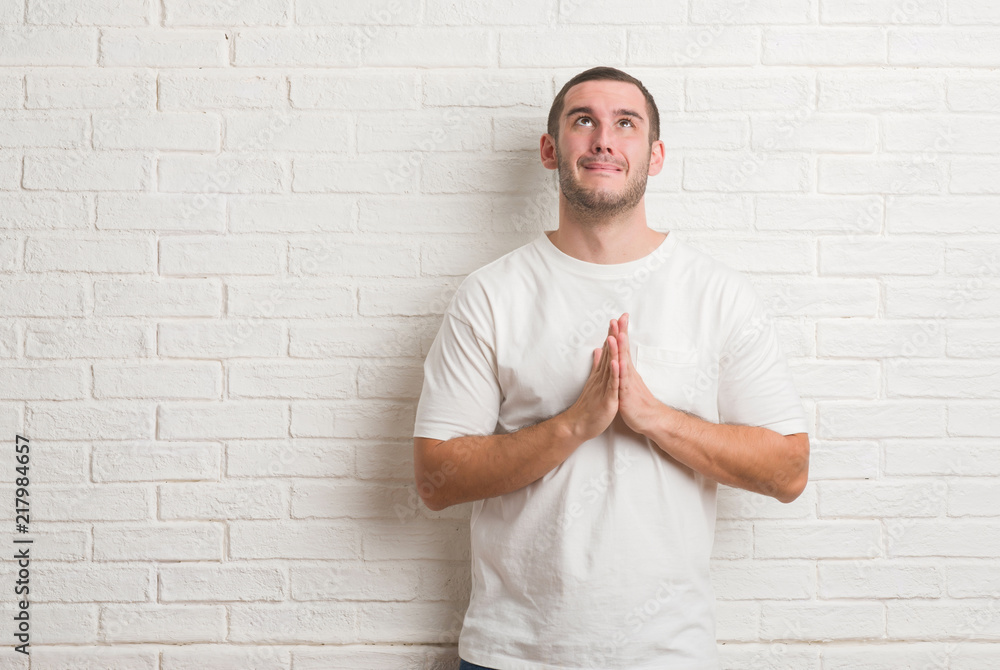 Young caucasian man standing over white brick wall cheerful with a smile of face pointing with hand and finger up to the side with happy and natural expression on face looking at the camera. Young caucasian man standing over white brick wall cheerful with a smile of face pointing with hand and finger up to the side with happy and natural expression on face looking at the camera.