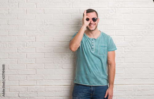 Young caucasian man standing over white brick wall wearing sunglasses with happy face smiling doing ok sign with hand on eye looking through fingers