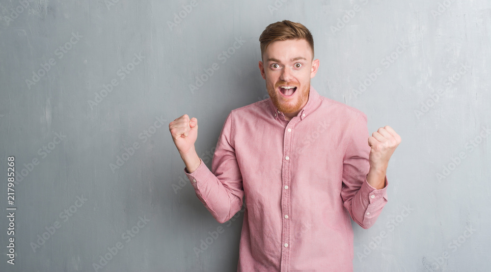 Young redhead man over grey grunge wall wearing pink shirt celebrating surprised and amazed for success with arms raised and open eyes. Winner concept.
