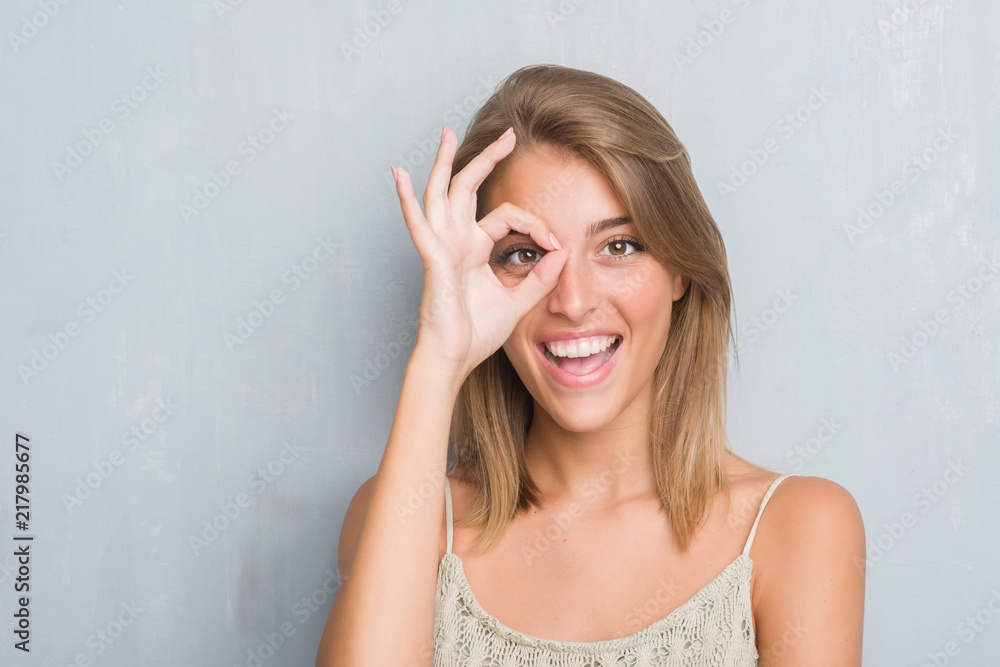 Beautiful young woman standing over grunge grey wall with happy face smiling doing ok sign with hand on eye looking through fingers Beautiful young woman standing over grunge grey wall with happy face smiling doing ok sign with hand on eye looking through fingers