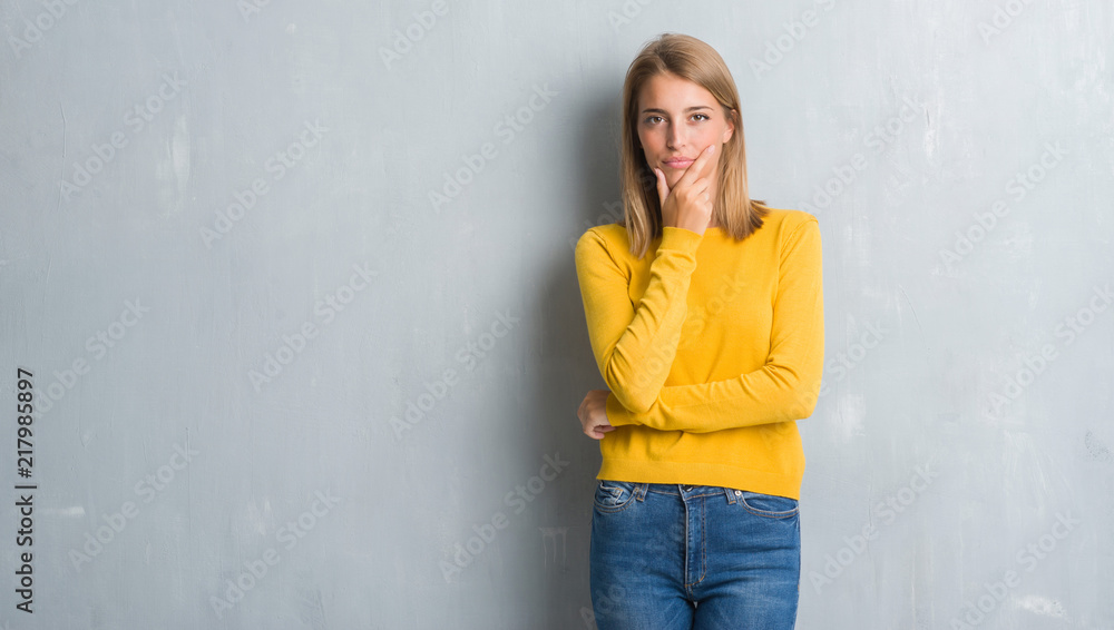 Beautiful young woman standing over grunge grey wall looking confident at the camera with smile with crossed arms and hand raised on chin. Thinking positive. Beautiful young woman standing over grunge grey wall looking confident at the camera with smile with crossed arms and hand raised on chin. Thinking positive.