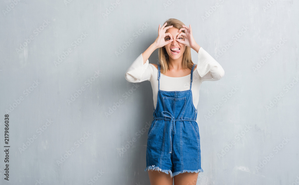 Beautiful young woman standing over grunge grey wall doing ok gesture like binoculars sticking tongue out, eyes looking through fingers. Crazy expression. Beautiful young woman standing over grunge grey wall doing ok gesture like binoculars sticking tongue out, eyes looking through fingers. Crazy expression.