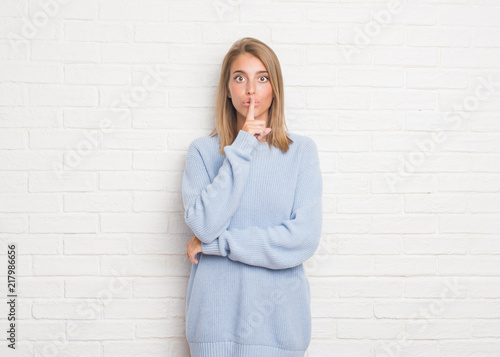 Beautiful young woman over white brick wall asking to be quiet with finger on lips. Silence and secret concept.