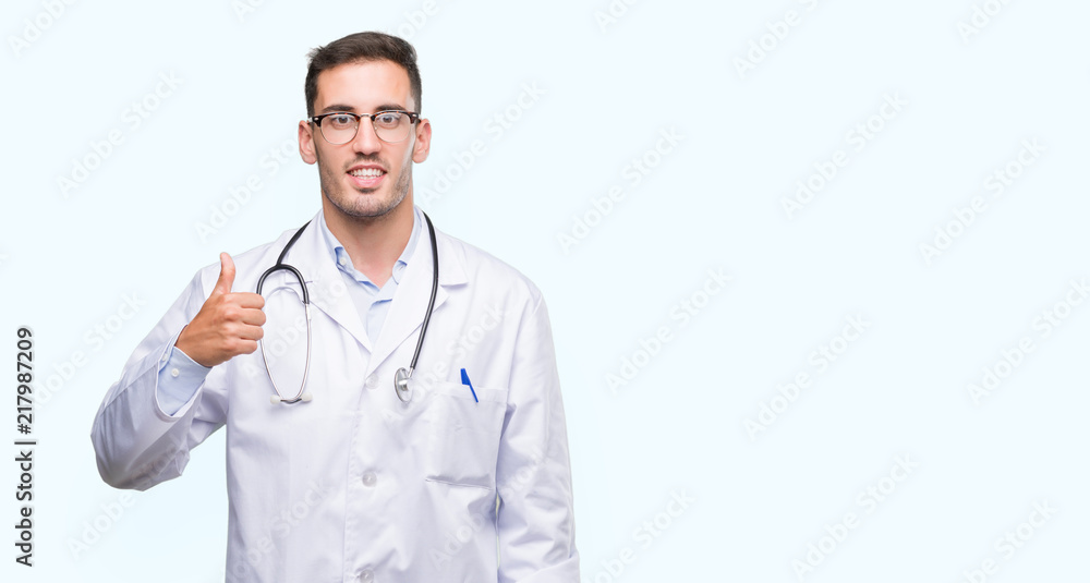 Handsome young doctor man doing happy thumbs up gesture with hand. Approving expression looking at the camera with showing success.