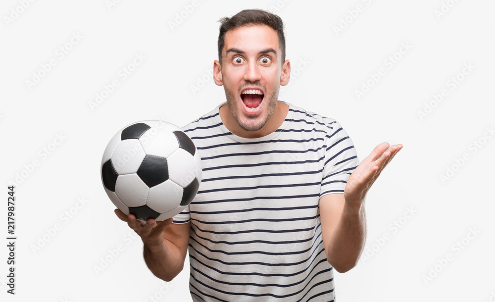 Handsome young man holding soccer football very happy and excited, winner expression celebrating victory screaming with big smile and raised hands Handsome young man holding soccer football very happy and excited, winner expression celebrating victory screaming with big smile and raised hands