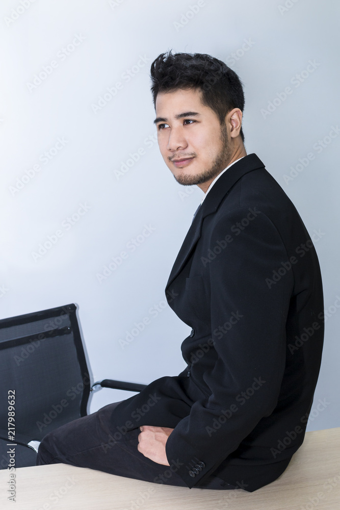 Young handsome businessman smiling and smart sit on table at office.He is 20-30 years old in black suit.Copy space. Young handsome businessman smiling and smart sit on table at office.He is 20-30 years old in black suit.Copy space.