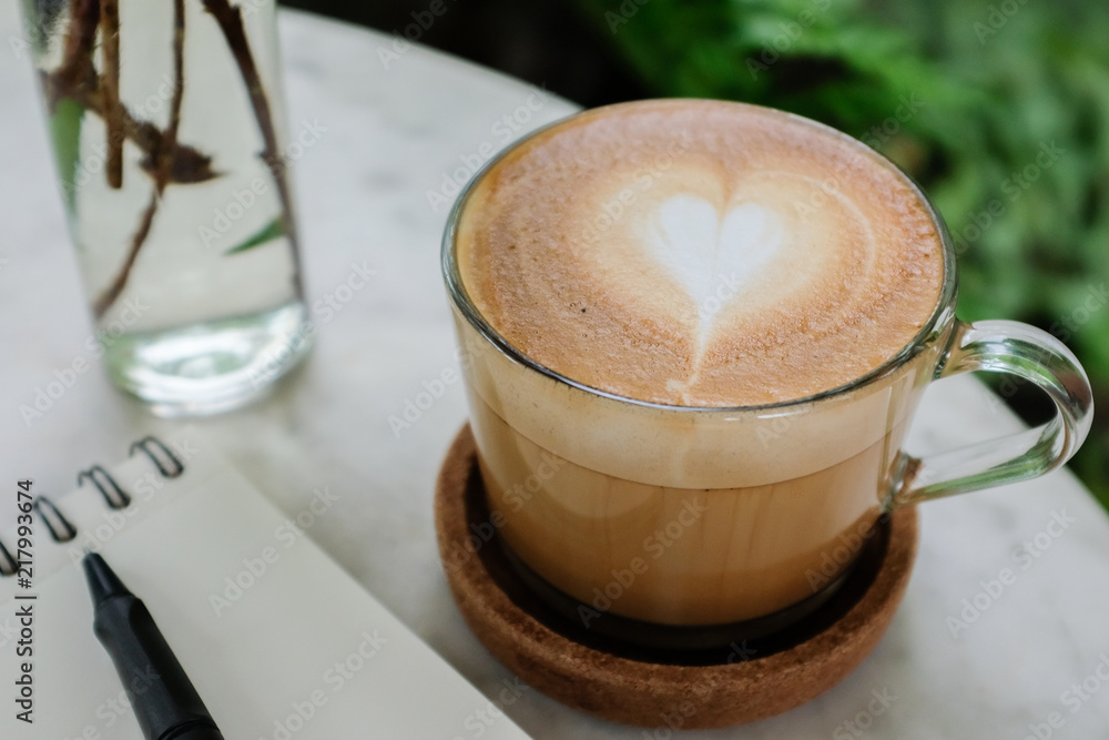 Close up caffe latte coffee in a glass cup on a marble coffee table with white notebook black pen glass vase with green background Close up caffe latte coffee in a glass cup on a marble coffee table with white notebook black pen glass vase with green background