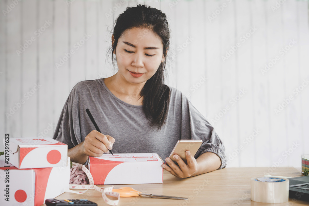 beautiful Asian woman selling online from home writing on box preparing order with smart phone in hand beautiful Asian woman selling online from home writing on box preparing order with smart phone in hand