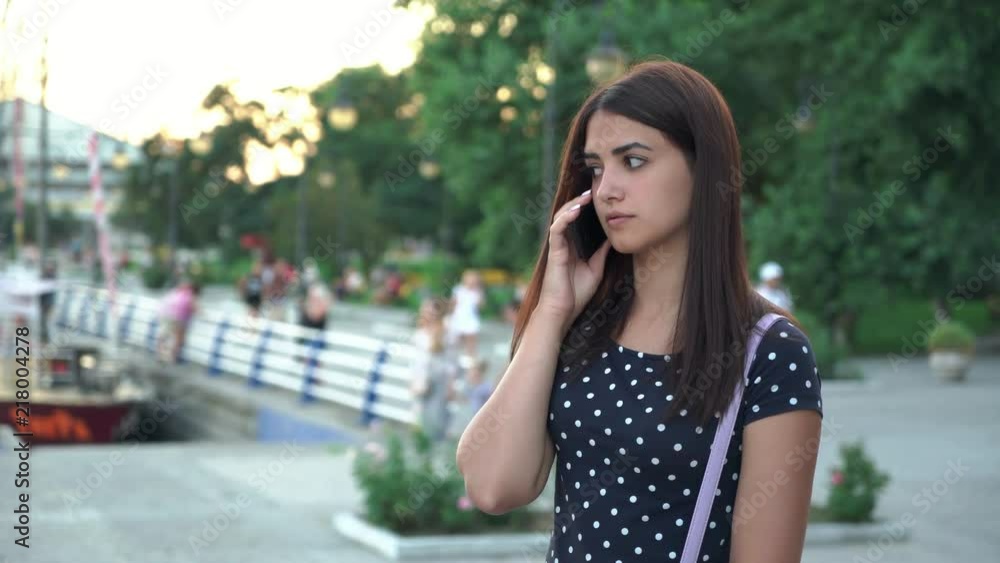 Portrait of a concerned beautiful young woman with sad eyes and long hair talking on phone with her relatives on a riverbank in summer Portrait of a concerned beautiful young woman with sad eyes and long hair talking on phone with her relatives on a riverbank in summer