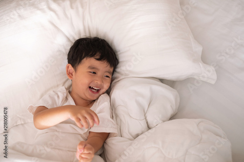 3 years old little cute Asian boy at home on the bed, kid lying playing and smiling on white bed with pillow and blanket, top view with copy space.