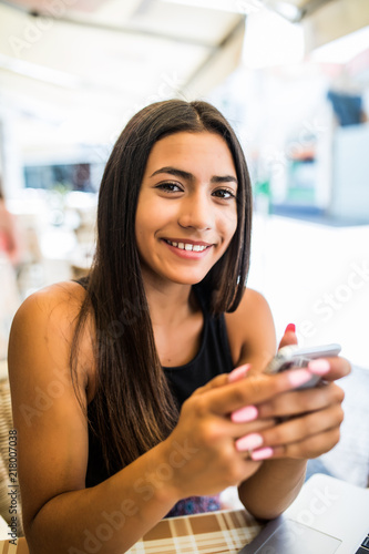 Smiling beautiful latin girl sitting in street cafe and texting message on her phone. Happy attractive woman browsing site on her smartphone with opened laptop in bar