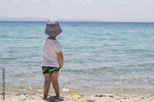 A little shy boy with hat standing in the sand near water and looking in the beautiful sea in front of him. Family trip and vacation concept. Summertime. photo