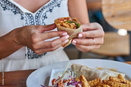 Female hands holding tasty mexican burrito with different ingredients inside. Woman eating delicious pita and salad with French fried potato.