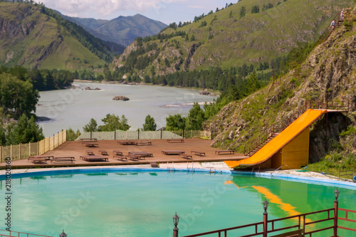 outdoor pool in the Altai mountains with a yellow water slide attraction with wooden chaise-longue, blue water, separated by a metal fence, overlooking the river, rocks and trees on a sunny day © Aleksandr Kondratov