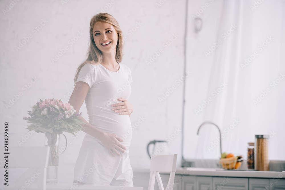 Fototapeta premium portrait of happy pregnant woman standing at table with bouquet of flowers in vase at home