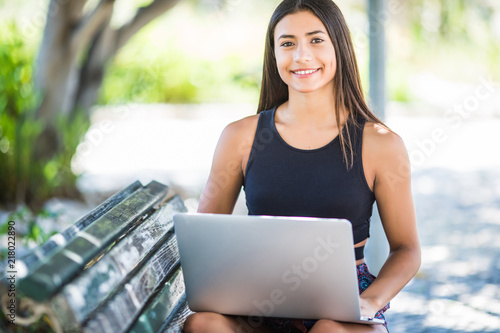 Pleased brunette latin woman isitting on bench in park and using laptop computer photo