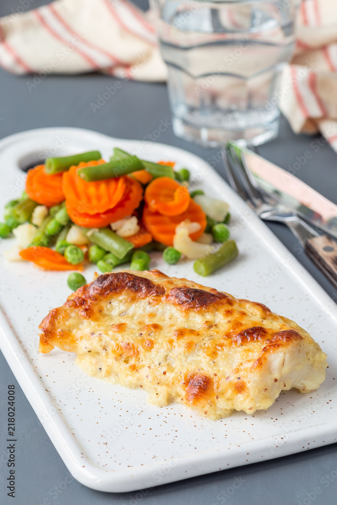 Baked cod fish fillet under cheese, mustard, pepper and cream crust, served with steamed vegetables, on ceramic board, vertical