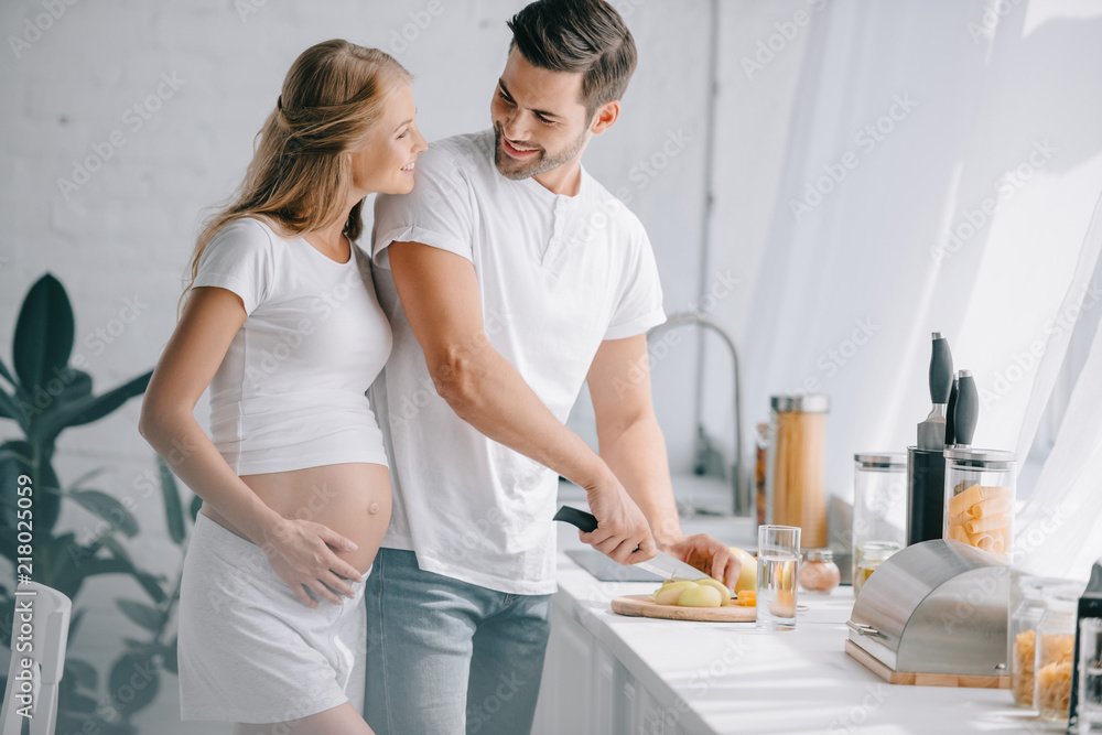 portrait of smiling man cutting fruits at counter with pregnant wife near by in kitchen at home portrait of smiling man cutting fruits at counter with pregnant wife near by in kitchen at home