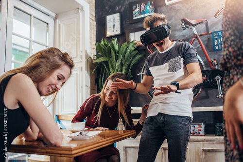 Young women laughing at their emotional male friend playing horror game in virtual reality goggles in cozy cafe photo