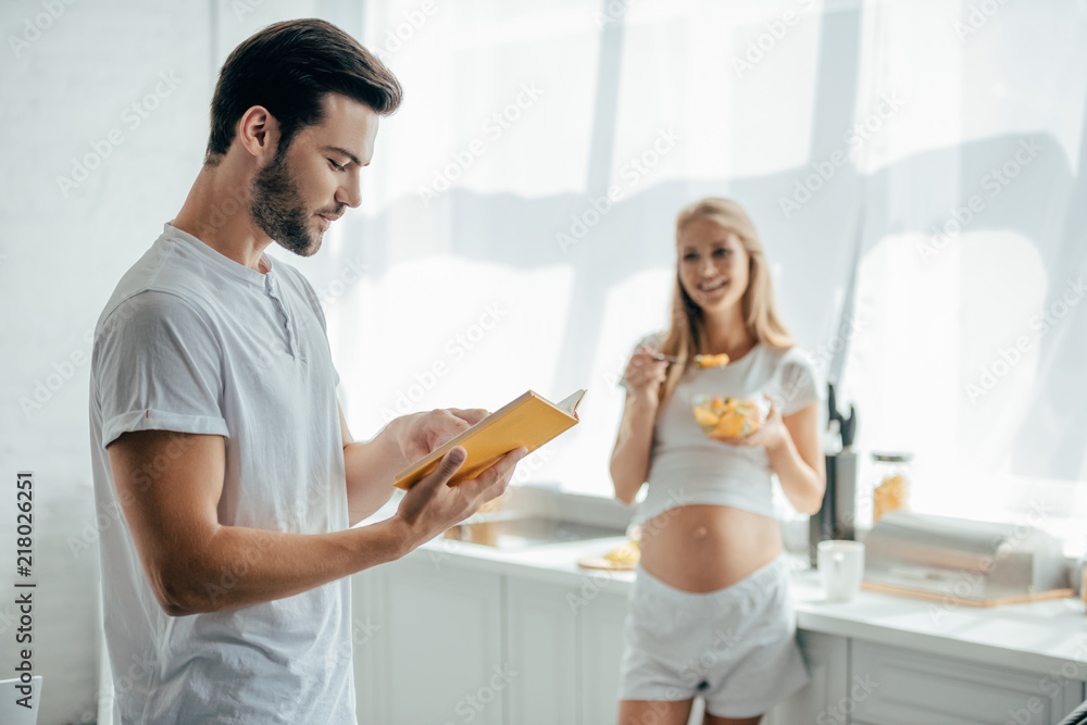 smiling pregnant wife with fruits salad and husband with book in kitchen at home smiling pregnant wife with fruits salad and husband with book in kitchen at home